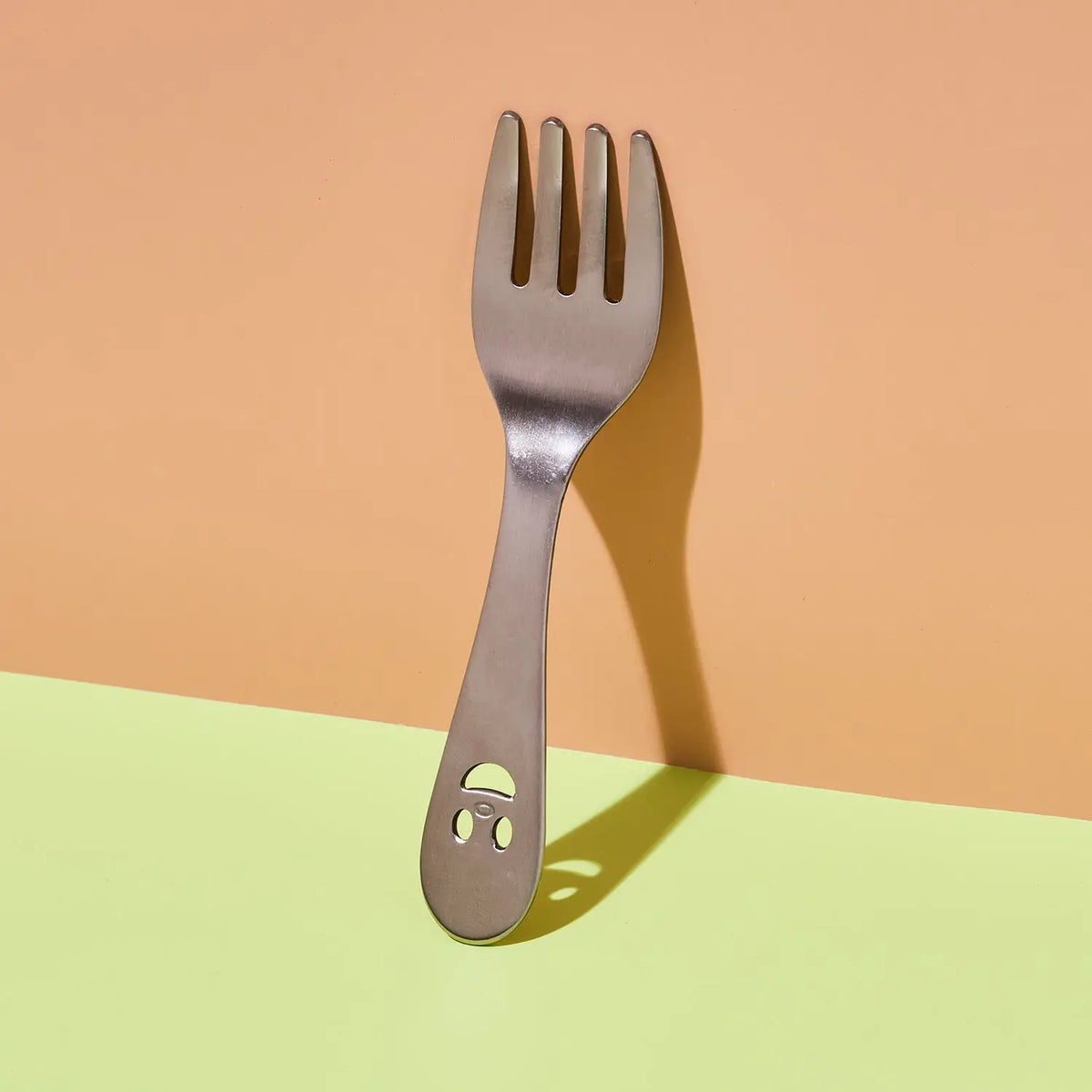 A stainless steel spork with a small smiley face cutout on the handle, standing upright against a two-tone peach and light green background.