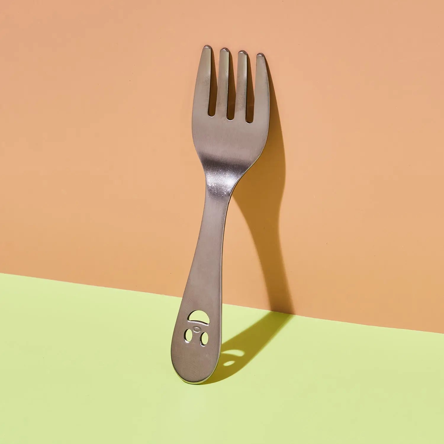 A stainless steel spork with a small smiley face cutout on the handle, standing upright against a two-tone peach and light green background.