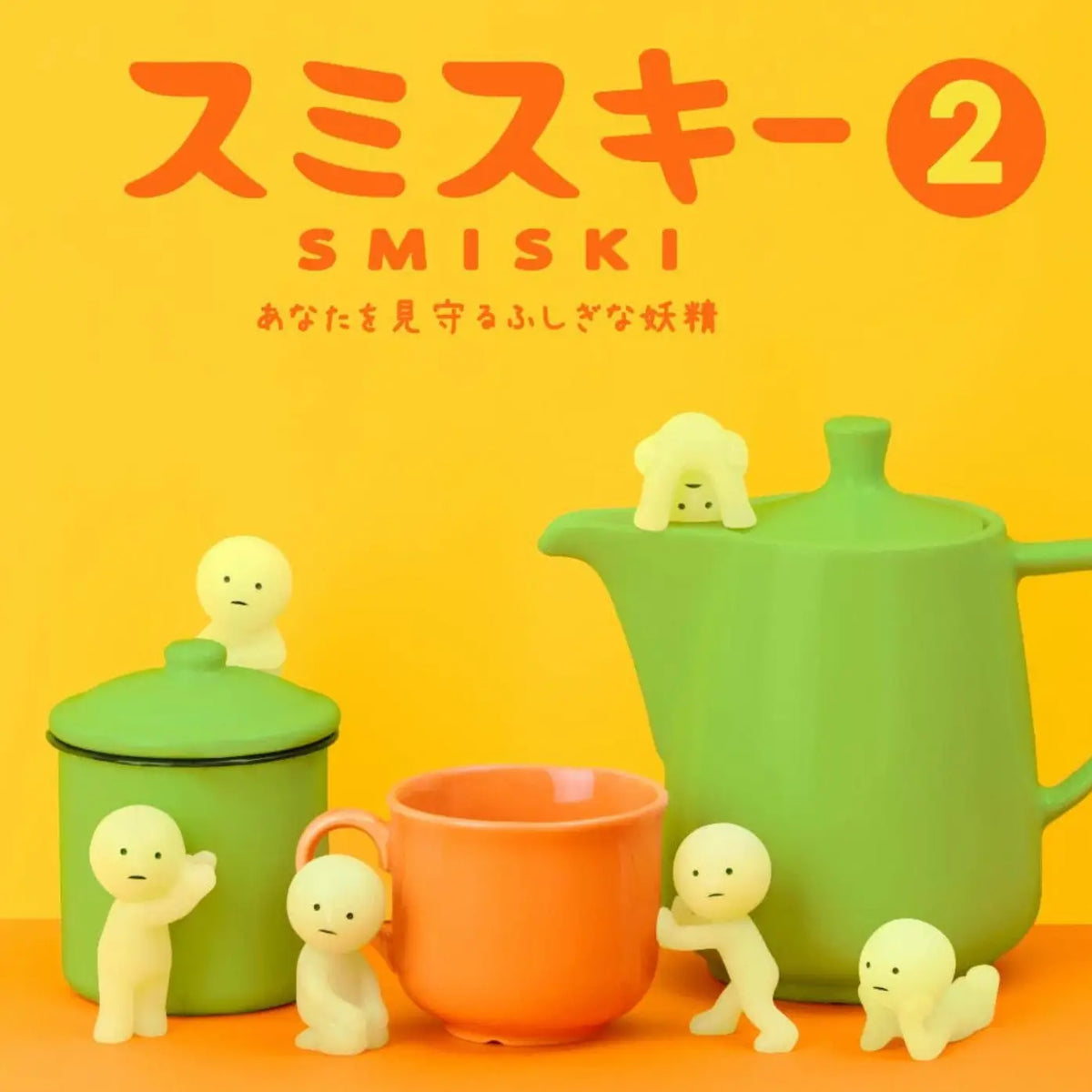 A set of pastel-colored tea-time accessories including a green teapot, a green lidded container, an orange mug, and small pale yellow cartoon figurines, arranged against a bright yellow and orange background with the text 'SMISKI' at the top.