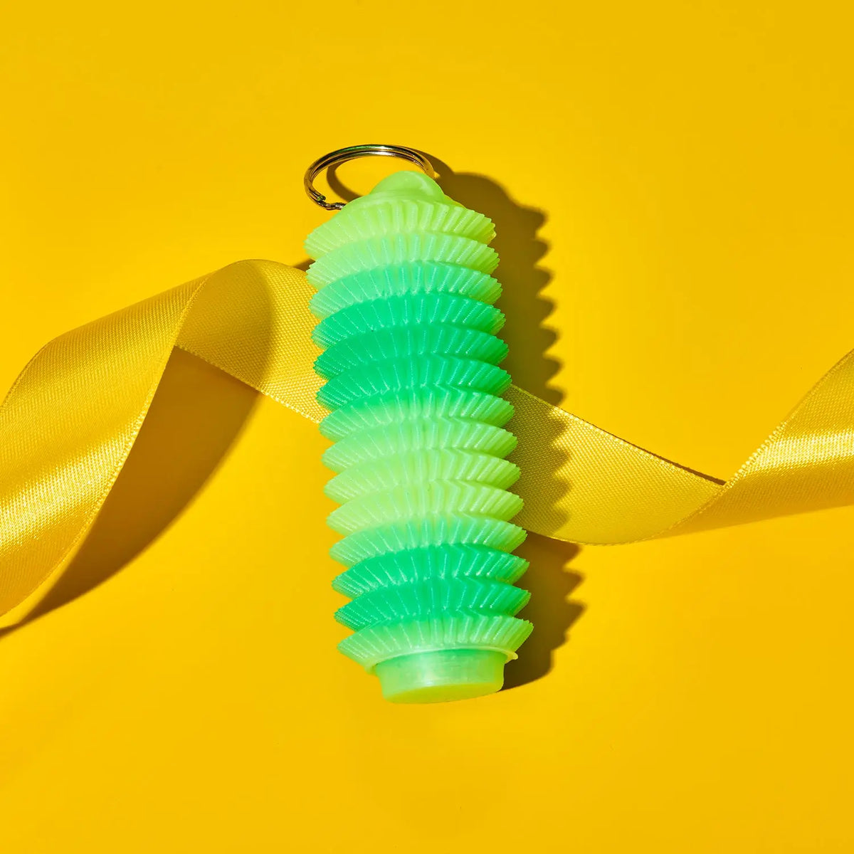 A green gradient fidget keychain holder with a gold metal keyring, wrapped in a yellow satin ribbon against a solid yellow background.