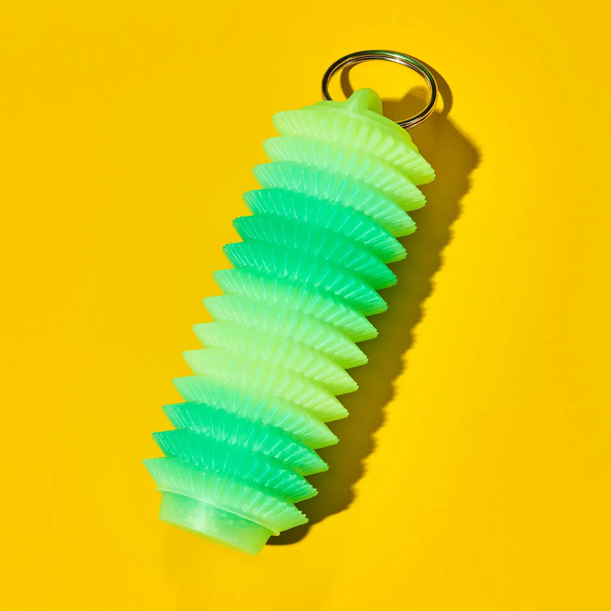 A green tube shaped fidget keychain with a gold metal key ring, shown on a bright yellow background.