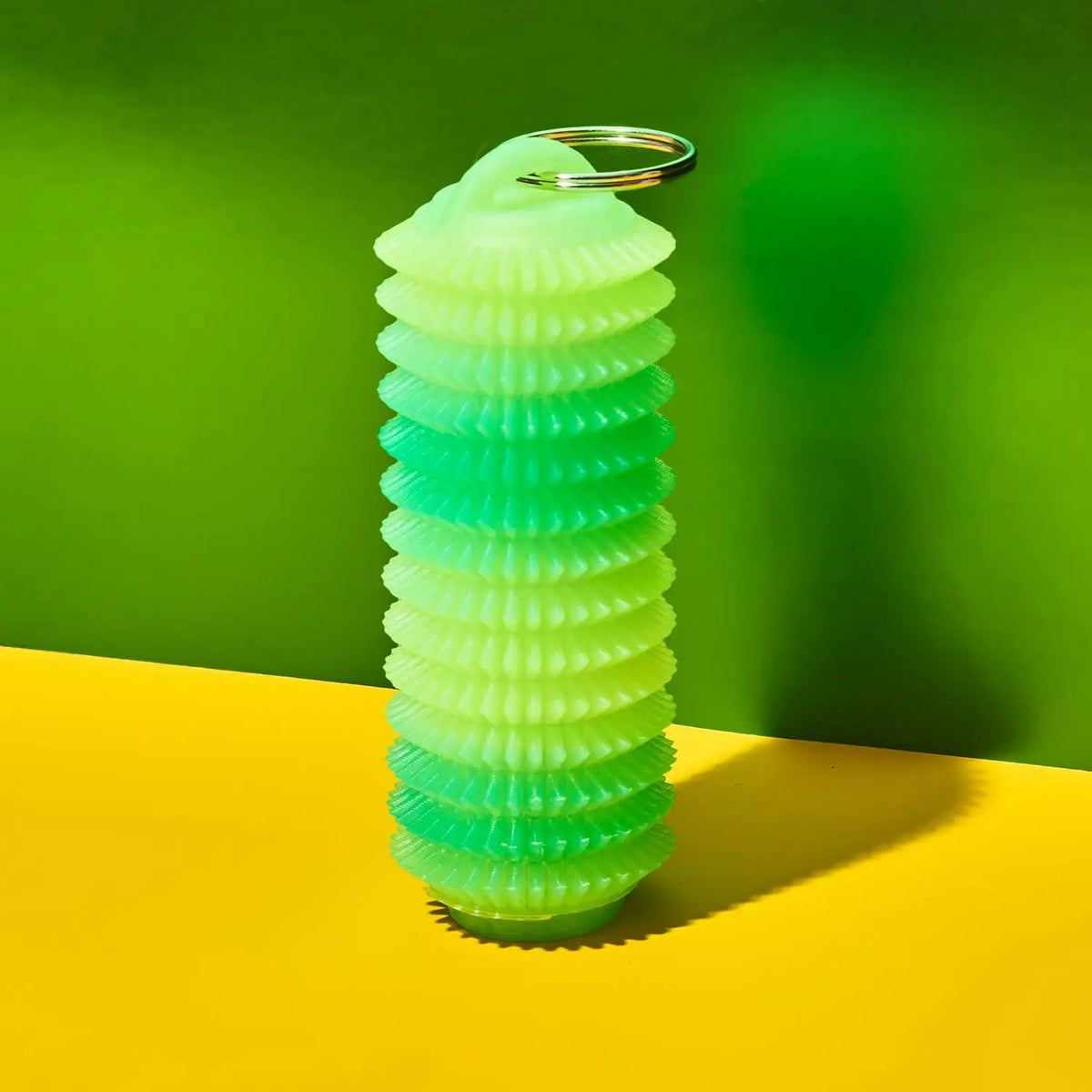 A translucent silicone keychain container shaped like a coiled spring, featuring a green-to-yellow gradient and a silver metal keyring at the top, set against a vibrant green and yellow background.