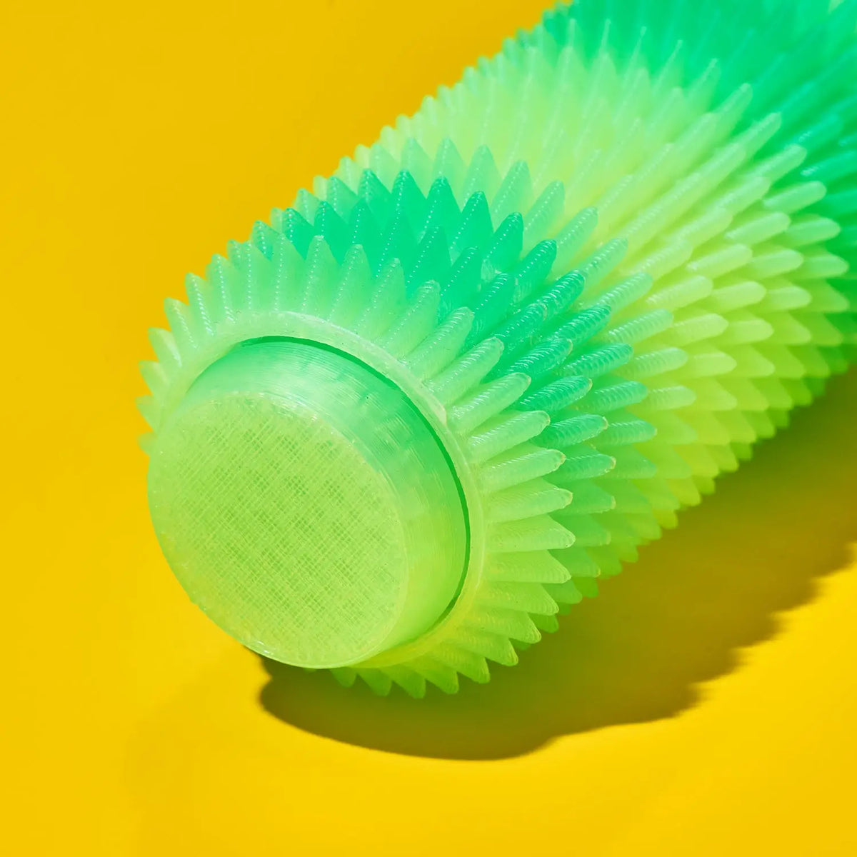 A close-up of a textured, ribbed cylindrical fidget key chain in a translucent neon green gradient, resting on a bright yellow background.
