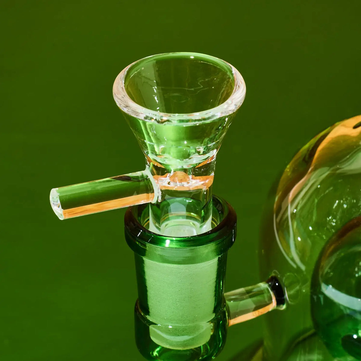 A close-up of a green glass bong component with a flared bowl and angled glass mouthpiece, set against a solid green background.