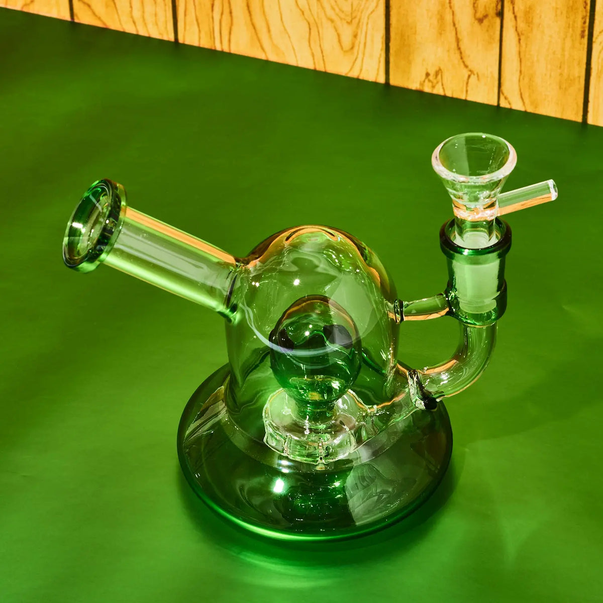A green glass water pipe with a round base, a curved mouthpiece, and a side attachment, photographed on a green surface with a wood panel background.