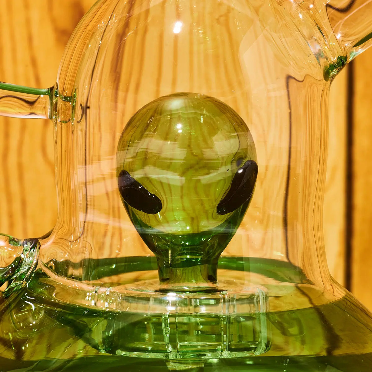 A close-up of a green glass water pipe green bulb-shaped alient in the center with dark eye-like shapes, set against a warm wood background.
