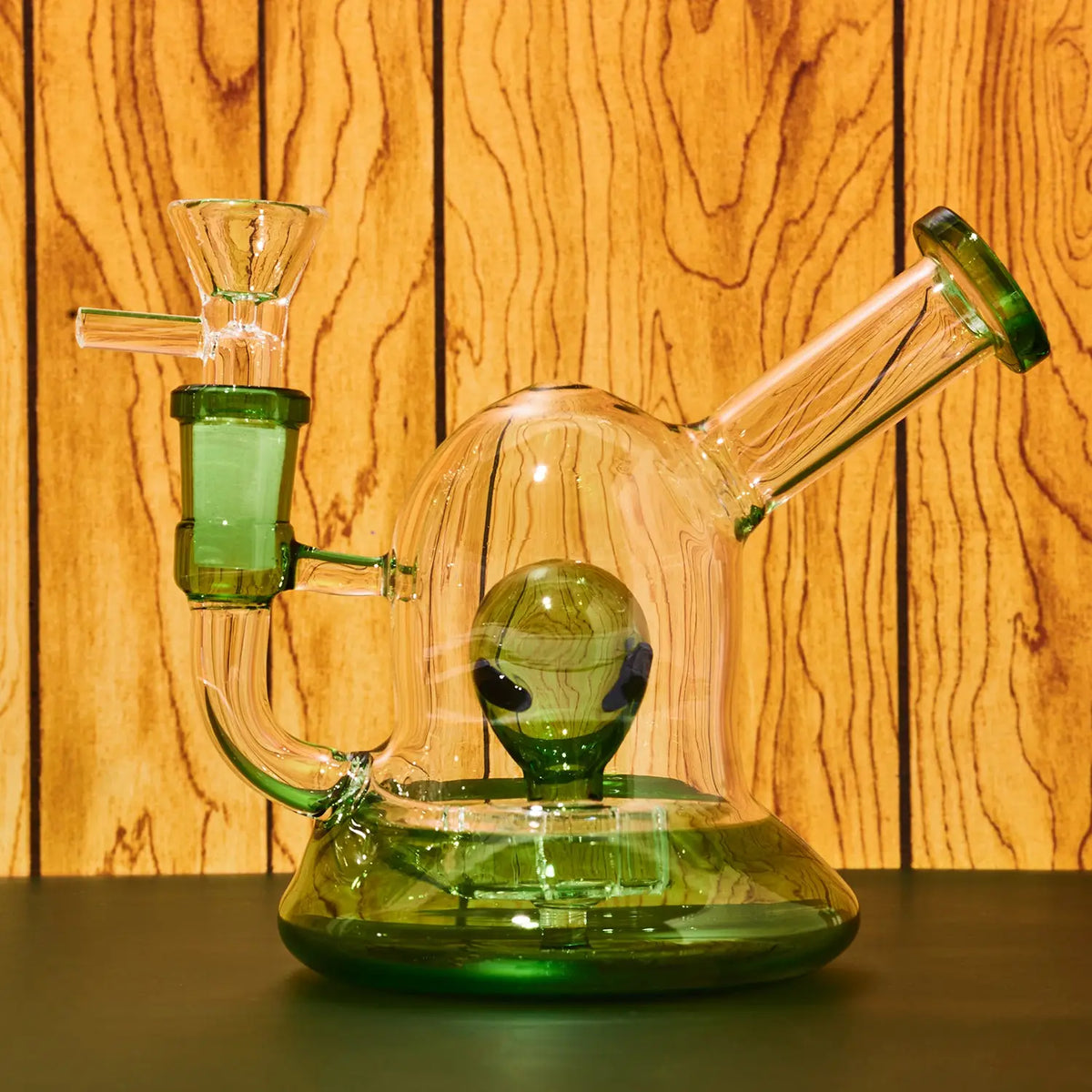 A green-tinted glass water pipe with a clear glass dome and a small alien figure inside, set against a warm wood-paneled background.
