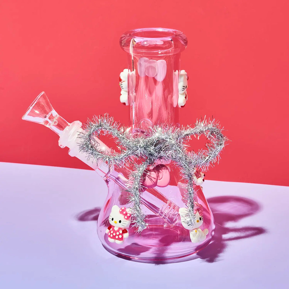 A pink glass water pipe with decorative white Hello Kitty figures and silver tinsel, set against a split red and lavender background.