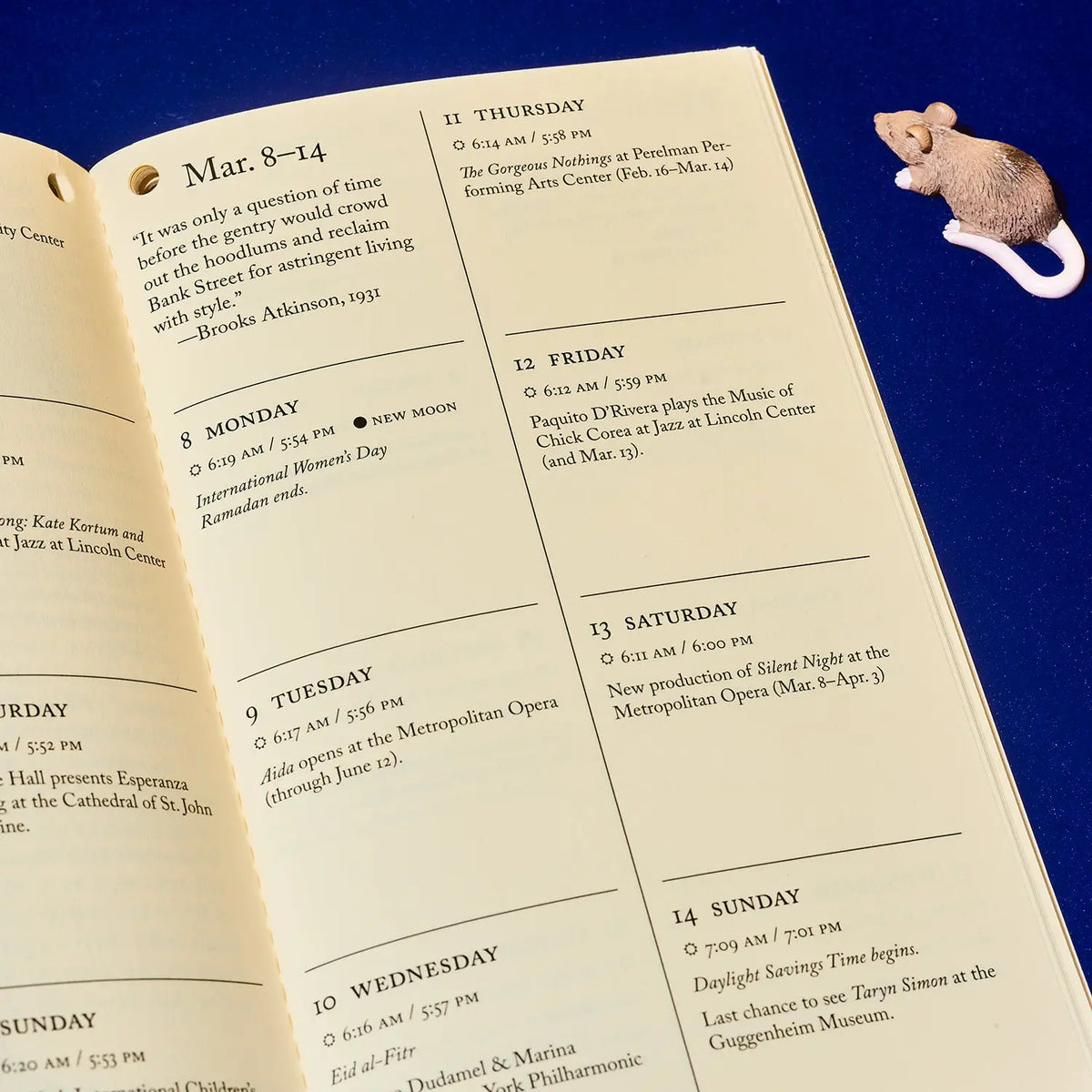An open printed weekly schedule or planner page with weekday blocks and notes, photographed on a dark blue background. A small illustrated mouse sits on the right edge of the page.