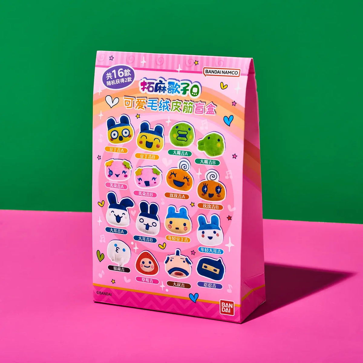 A pink paper bag package of Bandai Namco 'Mochi Dango Q' collectible blind boxes, standing on a pink surface against a green background.