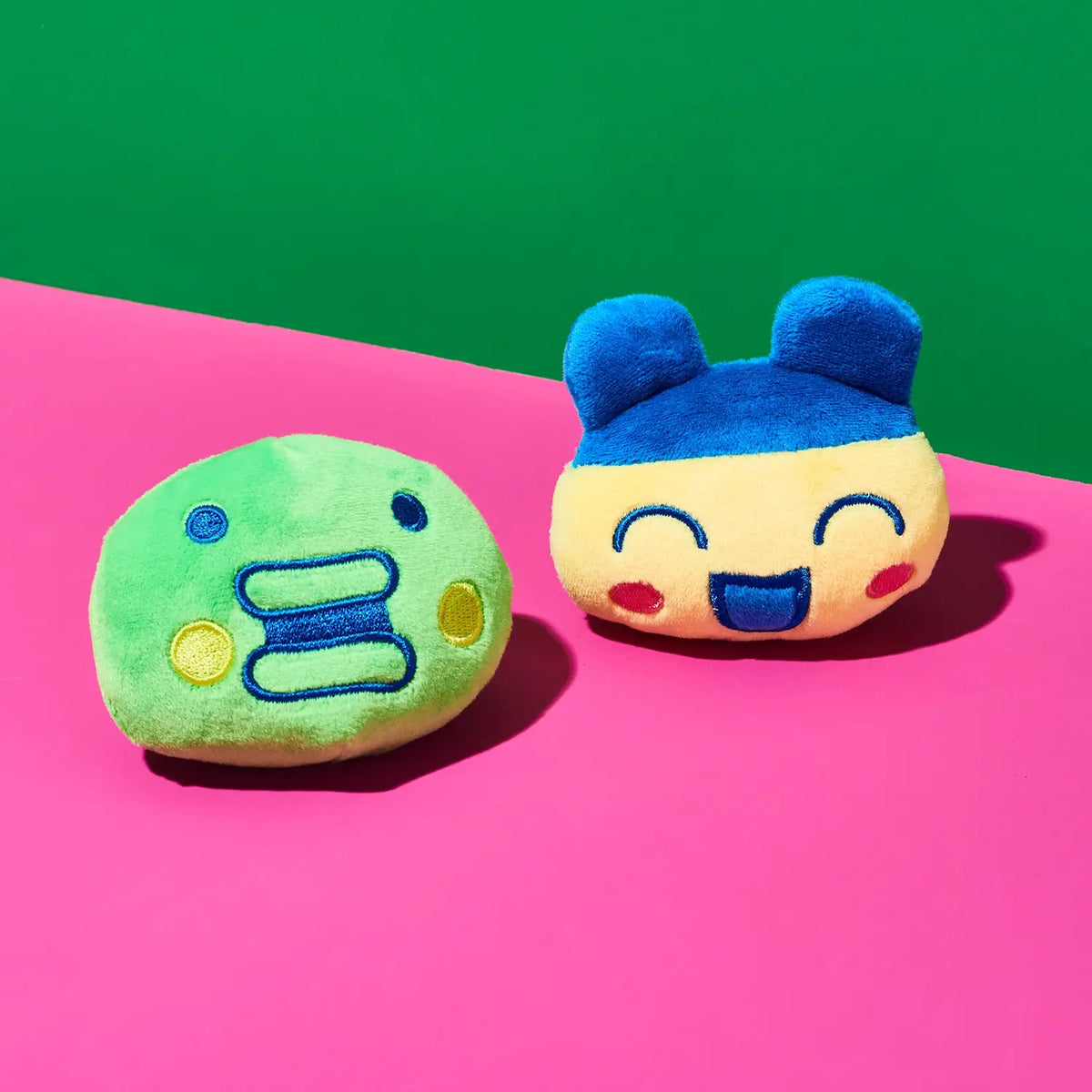 Two small plush character cushions on a bright pink surface with a green background. One cushion is light green with blue and yellow embroidered facial features.
