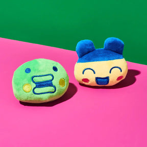 Two small plush character cushions on a bright pink surface with a green background. One cushion is light green with blue and yellow embroidered facial features.