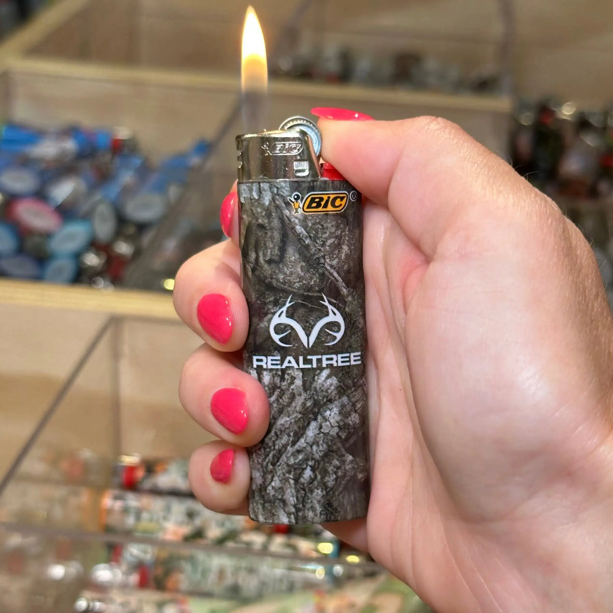 A hand with pink nail polish holds a BIC Realtree lighter with a dark gray and black camouflage pattern, lit and showing a flame, in front of a blurred retail display.