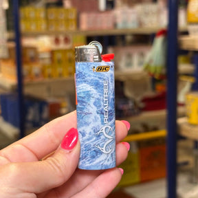 A hand with pink nail polish holds a BIC Realtree lighter featuring a blue and white marble wave pattern, set against a blurred background of a store shelf.