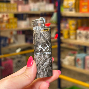 A hand with pink nail polish holds a BIC Realtree lighter with a gray and black camouflage pattern, set against a blurred background of a retail store shelf.