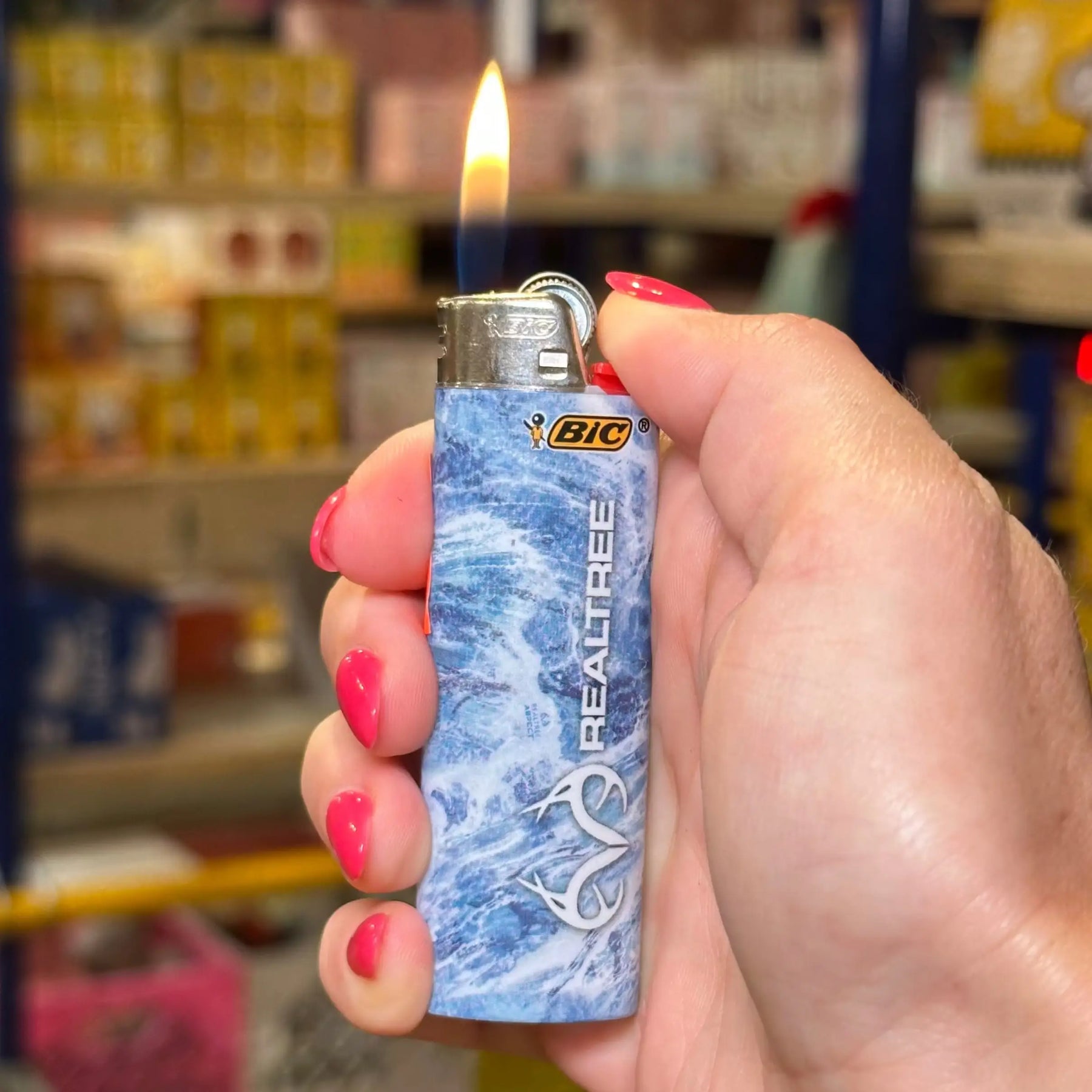 A hand with pink nail polish holds a lit BIC Realtree lighter against a blurred store background.