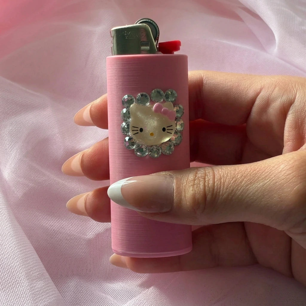 A pink lighter with a Hello Kitty face design surrounded by rhinestones, held in a hand against a soft pink fabric background.
