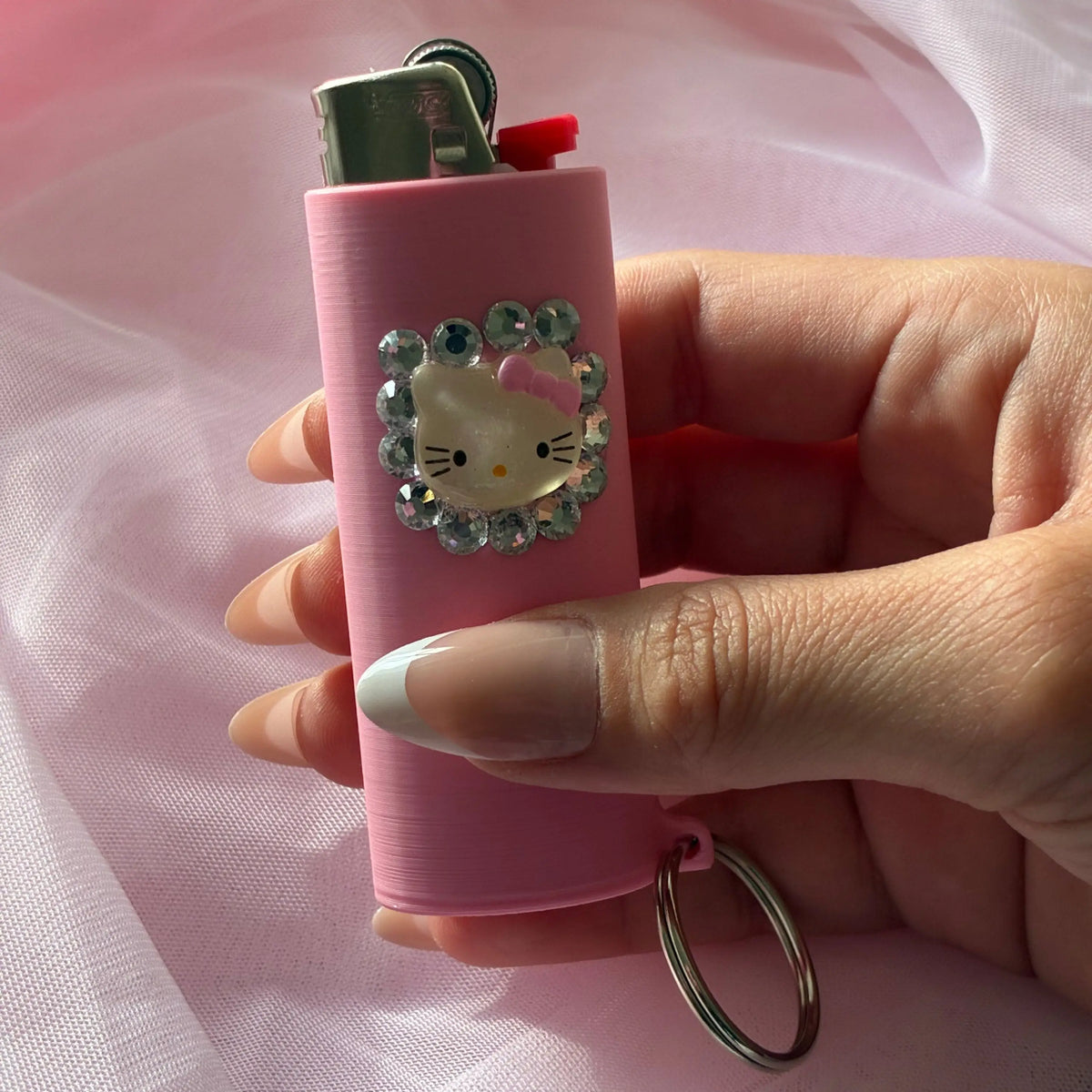 A pink lighter with a Hello Kitty face design surrounded by rhinestones, held in a hand against a soft pink fabric background.