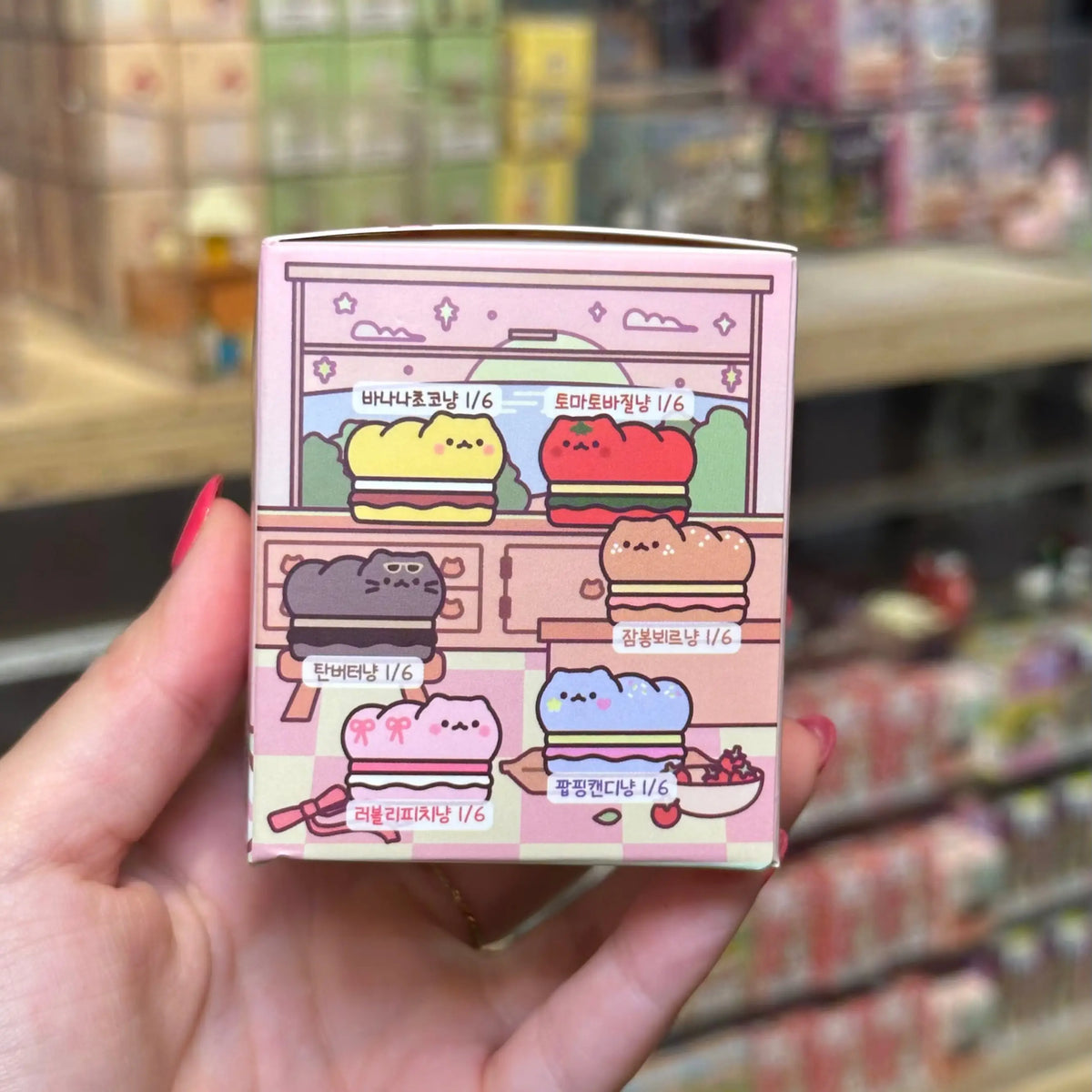 A hand with pink nail polish holds a small illustrated blind-box package featuring cute pastel character designs and Korean text, with a blurred store shelf background.