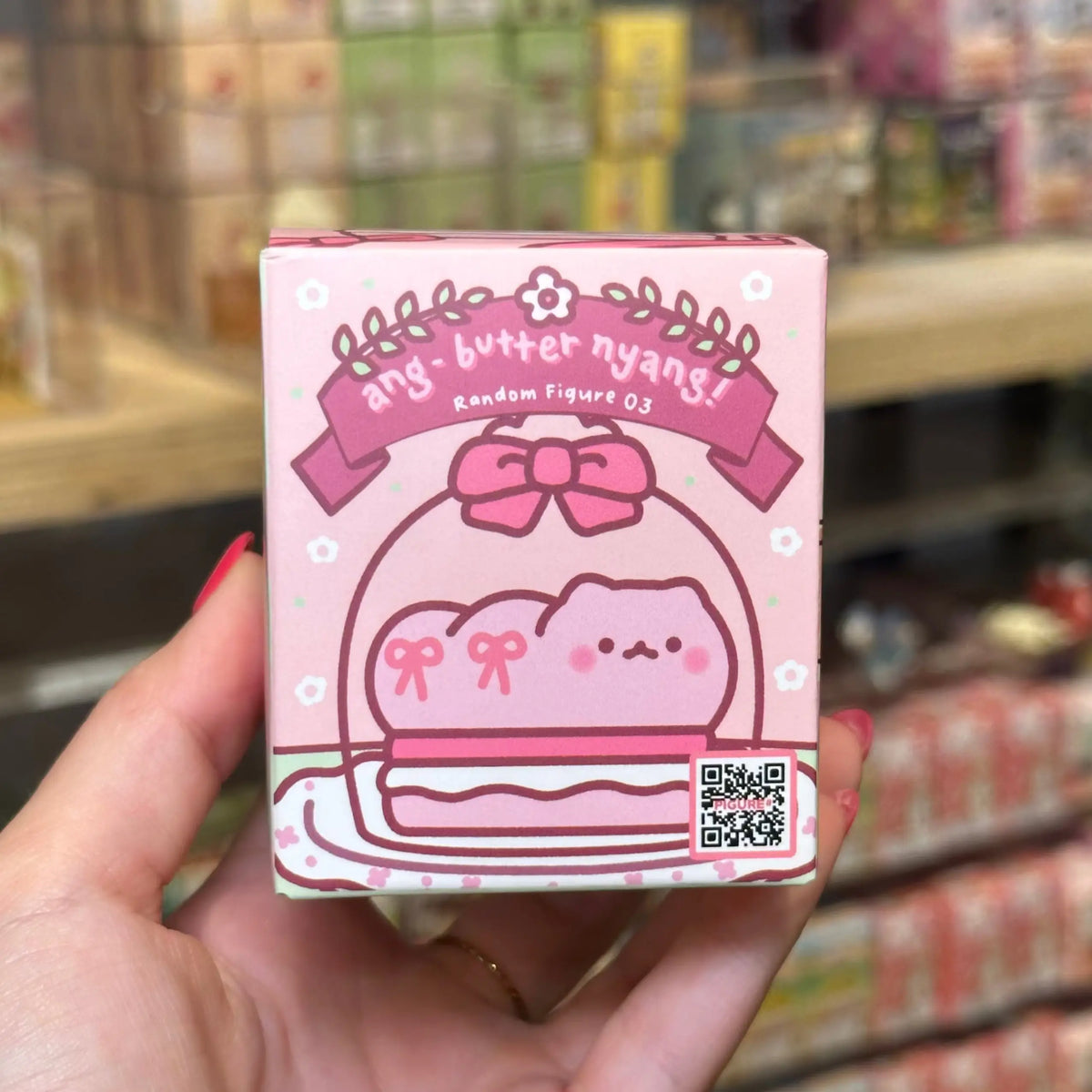 A hand holding a pink blind box for 'ang butter nyany' random figure 03, with a blurred store shelf background.