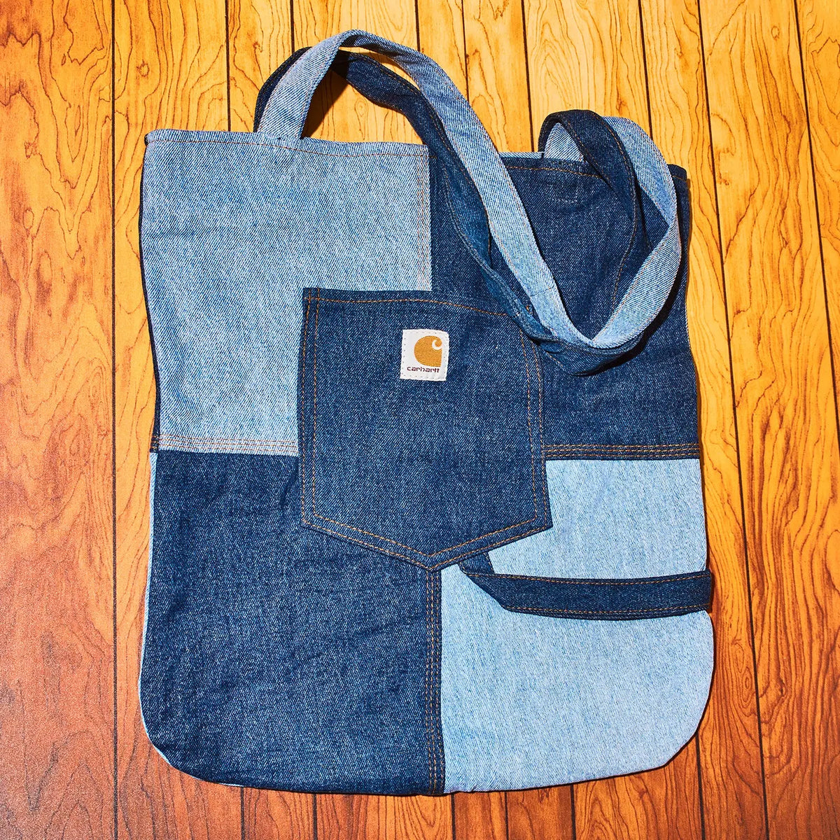 A patchwork denim tote bag made from light and dark blue denim panels, featuring a front pocket with a visible Carhartt logo patch and matching denim shoulder straps, laid flat on a wooden floor.