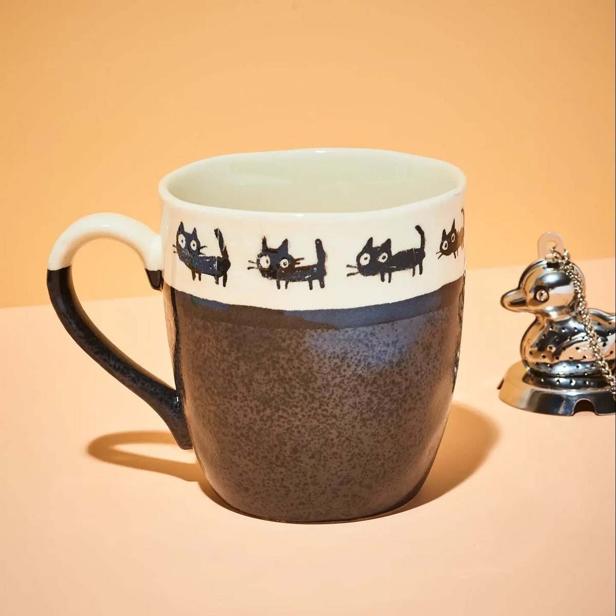 A ceramic mug with a speckled dark brown lower half, a white upper band decorated with black cat illustrations, and a black handle, shown on a peach-colored background next to a small silver duck figurine.