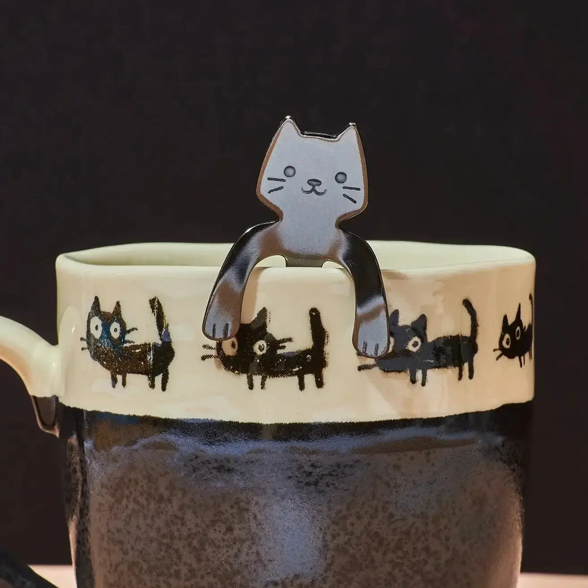 A ceramic mug with a dark speckled lower half and a cream upper band decorated with black cat illustrations. A metal spoon handle shaped like a white cat with black outlines rests on the rim of the mug against a dark background.