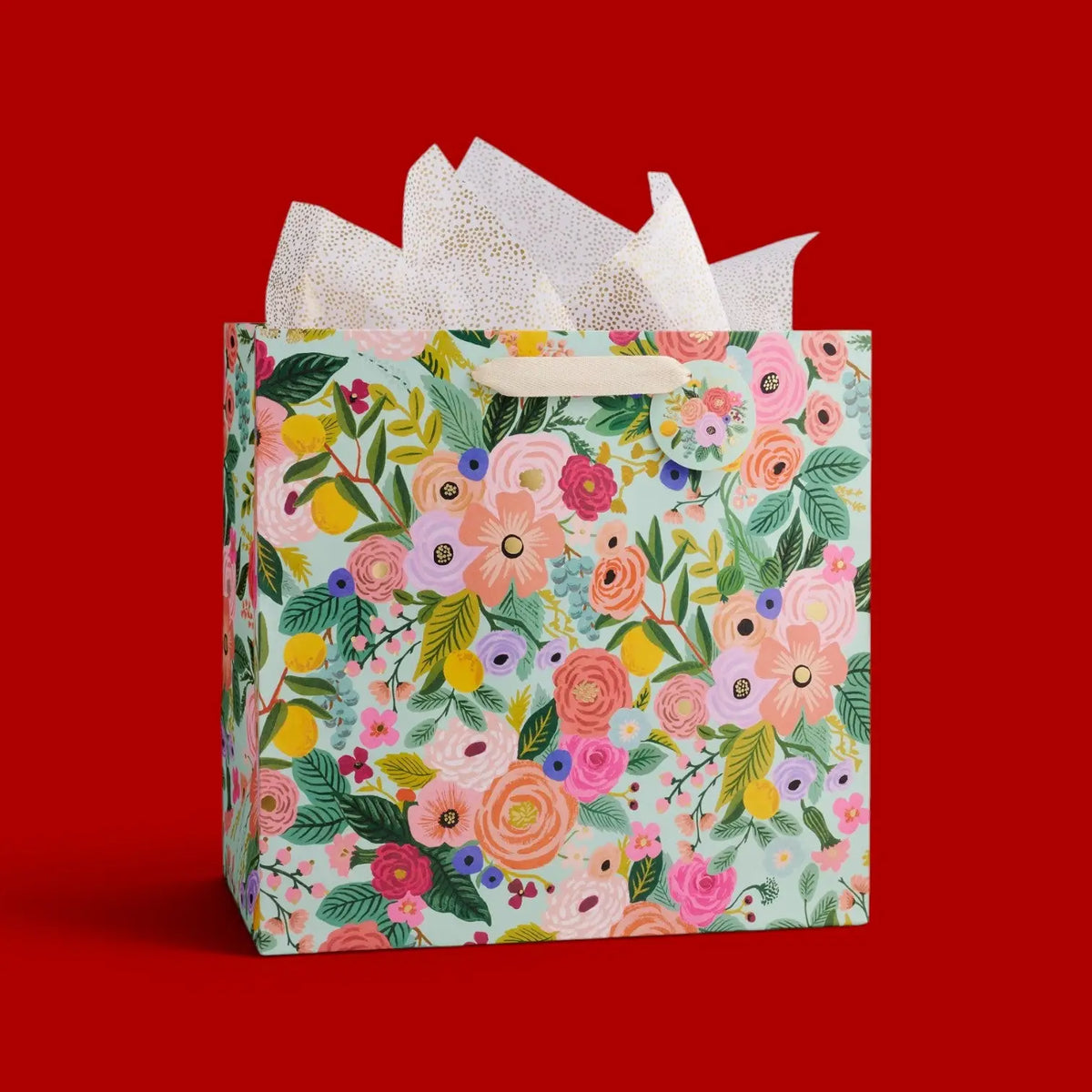 A square gift bag with a colorful floral print on a mint green background, filled with white tissue paper and set against a solid red background.