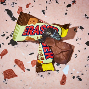 A close-up of a partially unwrapped Snickers ERASED chocolate bar, showing the brown chocolate coating and the cream-colored wrapper with red and blue lettering, placed on a pink terrazzo surface.