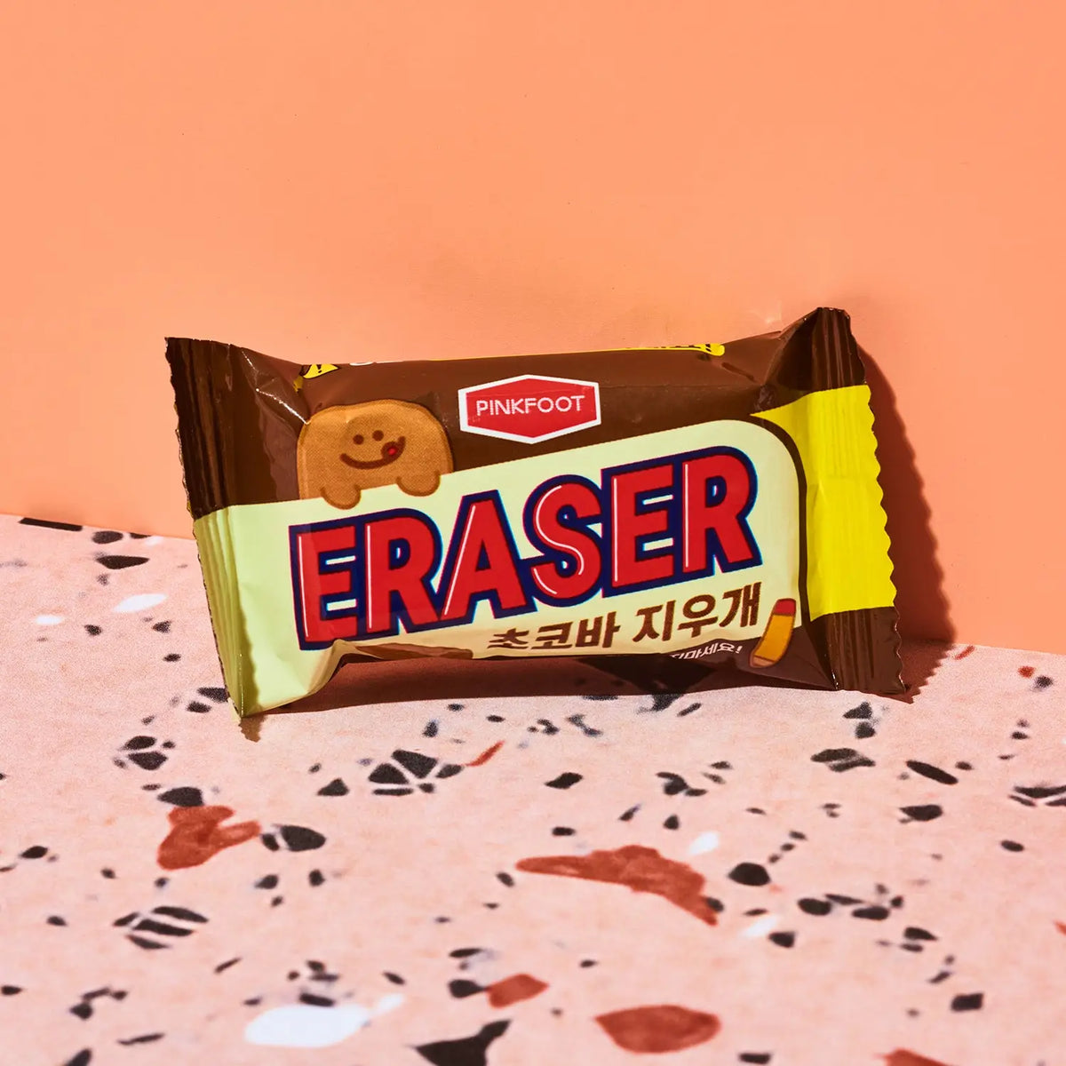 A packaged Pinkfoot Eraser cookie snack, wrapped in brown and yellow packaging with bold red and blue lettering, set on a terrazzo-patterned surface against a solid peach background.