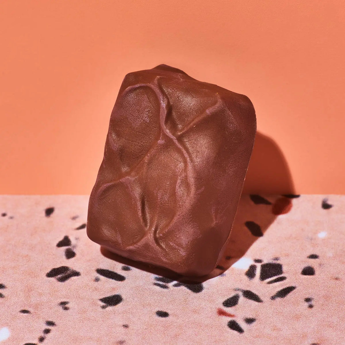 A single chocolate-covered rectangular product with a textured, crumpled surface stands upright on a speckled terrazzo-like surface against a solid peach-orange background.