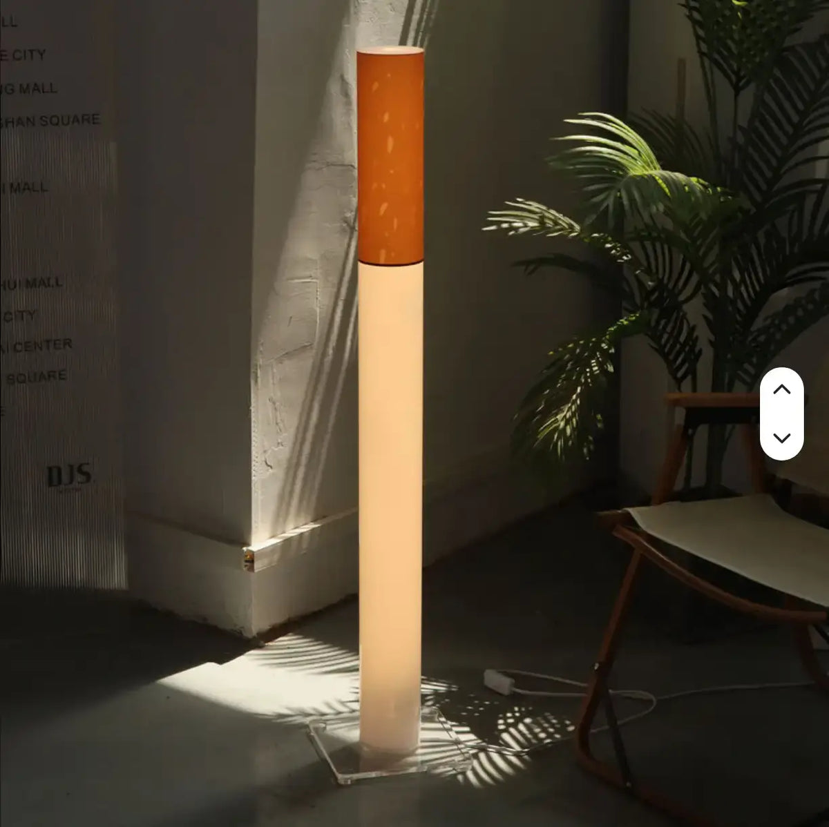 A modern floor lamp with a glowing white cylindrical body and a textured orange top section, standing in a styled room corner with a chair and plant nearby.