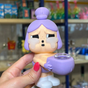 A hand with red nail polish holds a purple and peach-colored crybaby collectible bong toy figure that is also a bong with a purple hair bun and a small purple cup, set against a blurred store background.
