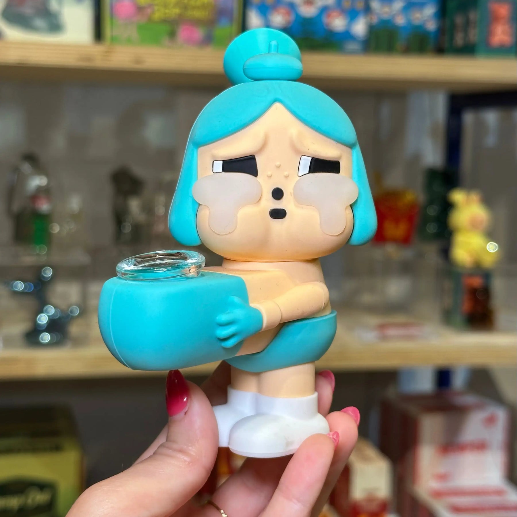 A hand holds a small cartoon-style collectible crybaby figure that is also bong with bright turquoise blue hair and matching turquoise accessories, standing in front of blurred shelves of boxed items.