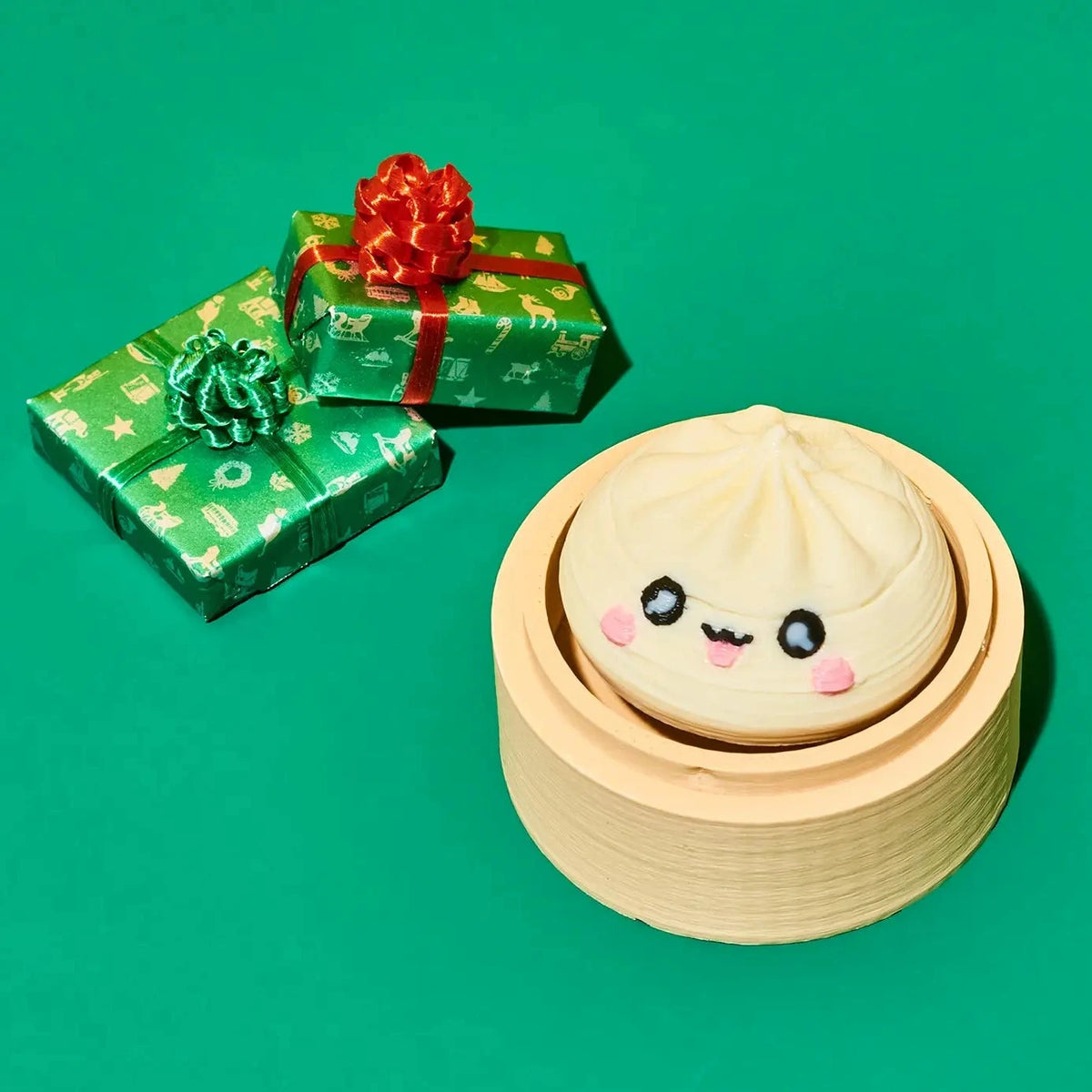 A cute, kawaii-style dumpling character with a smiling face sits inside a round bamboo steamer basket on a solid green background. Two wrapped gift boxes with green patterned paper and red bows are placed in the background.