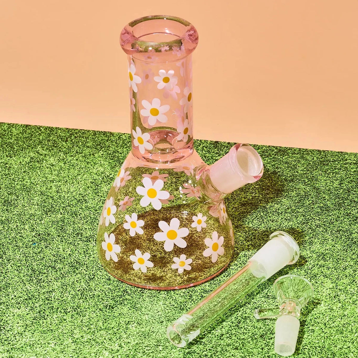 A pink glass beaker-style water pipe with a white and yellow daisy pattern, shown with two clear glass accessories on a green artificial turf surface against a peach background.