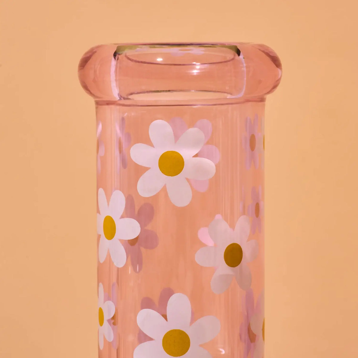 A close-up of a translucent pink glass container or vase decorated with a pattern of white daisies with yellow centers, set against a solid peach-colored background.