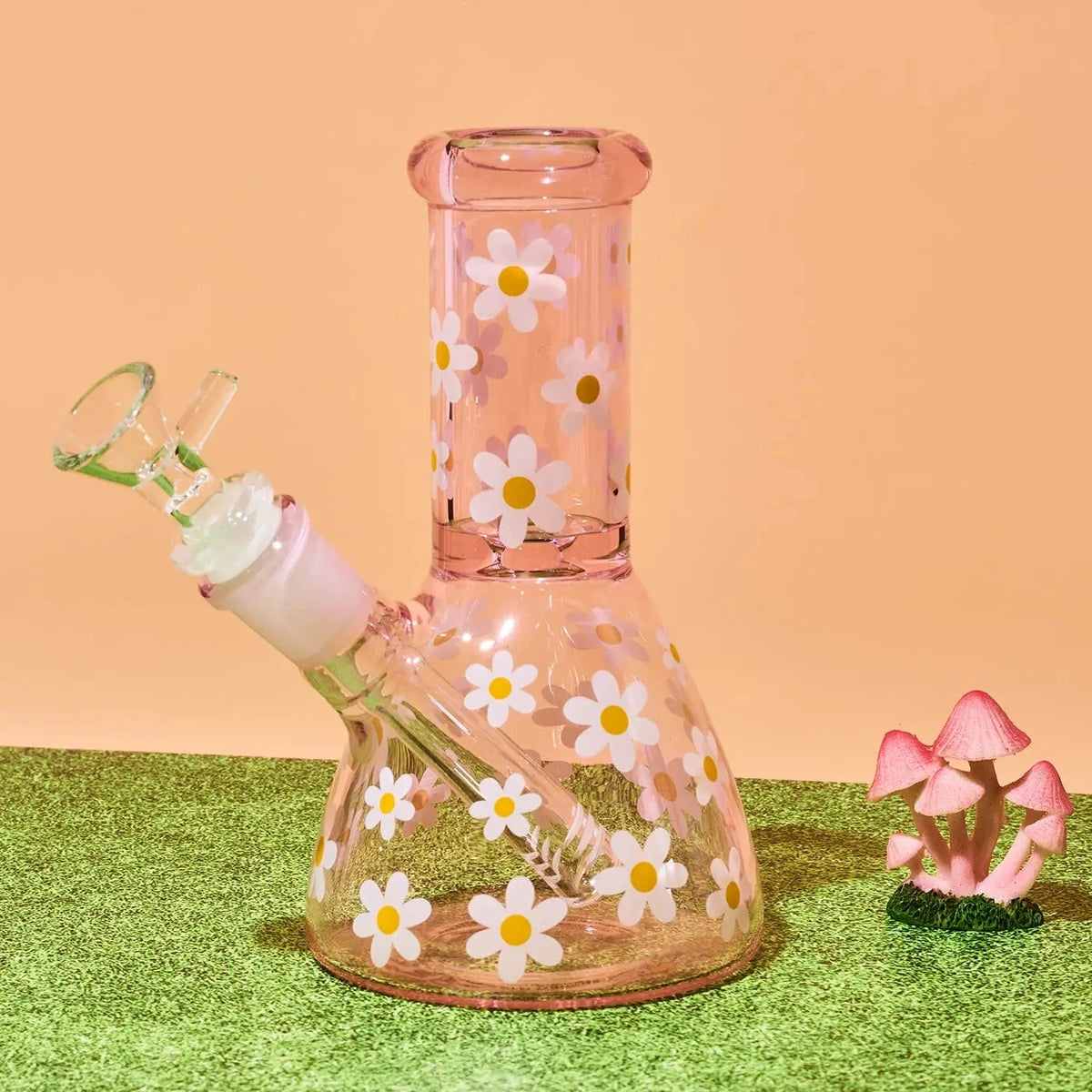 A pink glass water pipe with a daisy pattern, set on artificial green grass next to small pink mushrooms against a peach background.