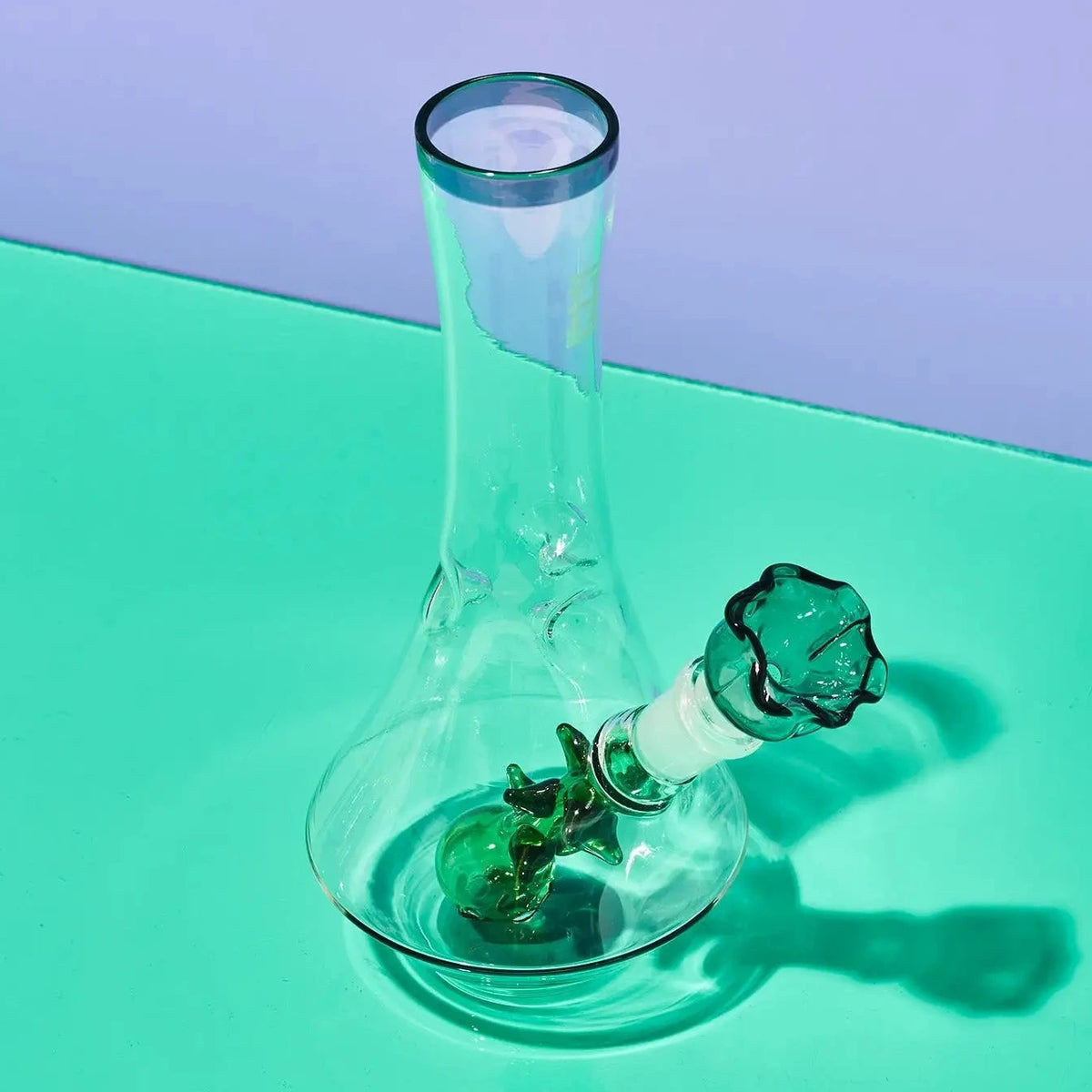 A clear glass water pipe with a green glass bowl and neck, set on a mint green surface against a lavender background.