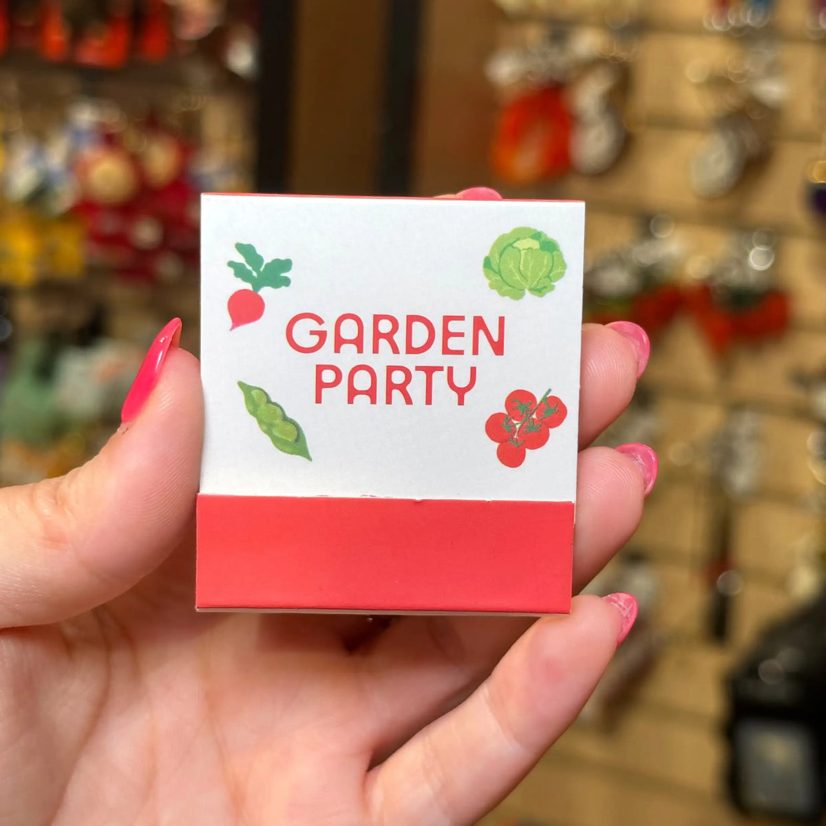 A hand with pink fingernails holds a small square box labeled 'GARDEN PARTY' with vegetable illustrations on a white top half and a coral-red bottom band. The background is a blurred retail store setting.