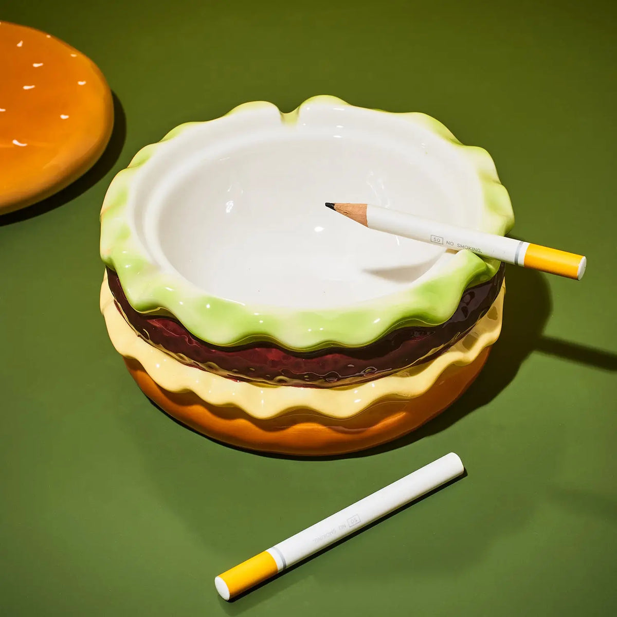 A novelty ceramic ashtray designed like a hamburger, featuring a white bowl, light green rind, brown patty, and yellow bun layers. Two toy cigarettes are placed on top and nearby, set against a solid green background.