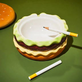 A novelty ceramic ashtray designed like a hamburger, featuring a white bowl, light green rind, brown patty, and yellow bun layers. Two toy cigarettes are placed on top and nearby, set against a solid green background.