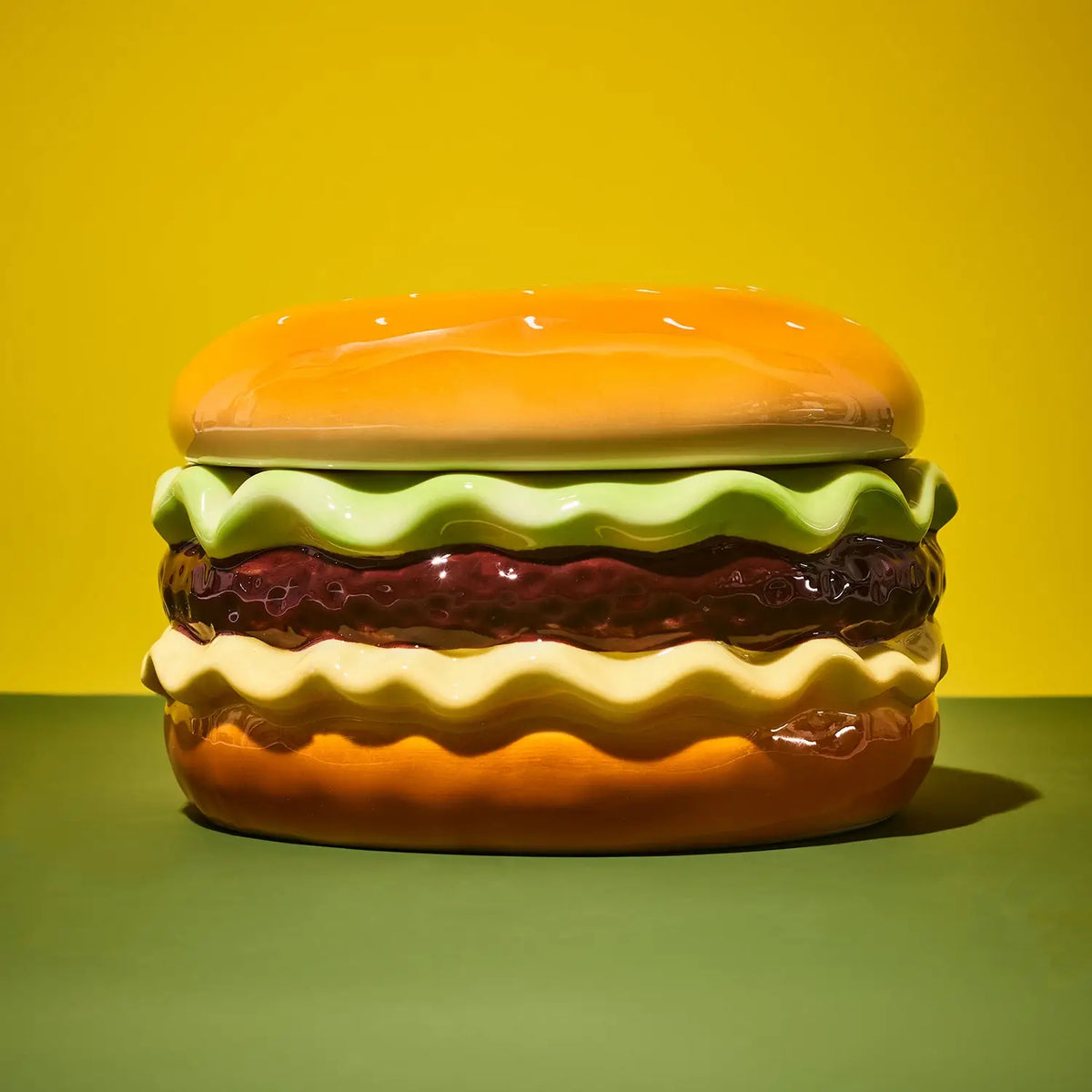 A glossy ceramic container shaped like a hamburger, featuring a yellow-orange bun, light green lettuce, dark red patty, and cream cheese, set against a yellow and green background.
