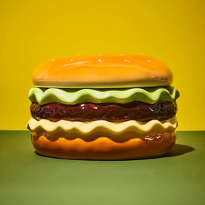 A glossy ceramic container shaped like a hamburger, featuring a yellow-orange bun, light green lettuce, dark red patty, and cream cheese, set against a yellow and green background.