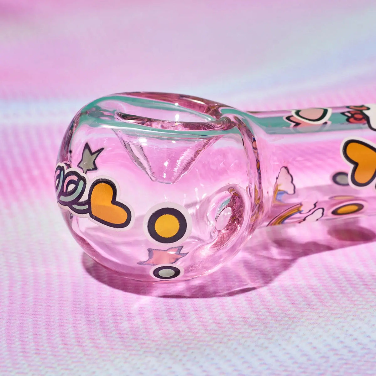 A clear glass water pipe with a pink tint and colorful cartoon-style illustrations, resting on a pink textured surface.