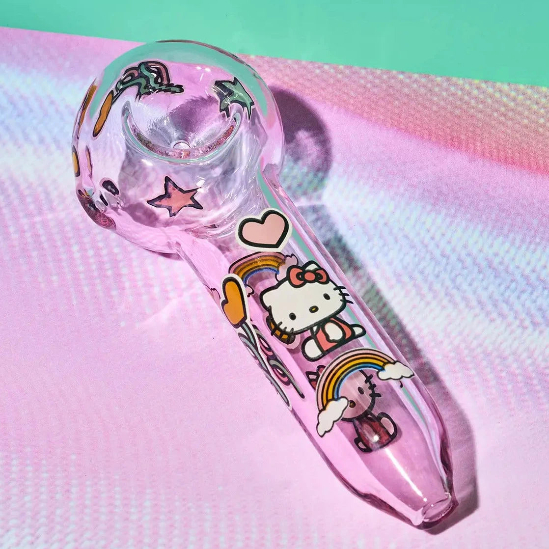 A pink glass water pipe decorated with Hello Kitty and rainbow illustrations, resting on a pink iridescent surface with a mint green background.