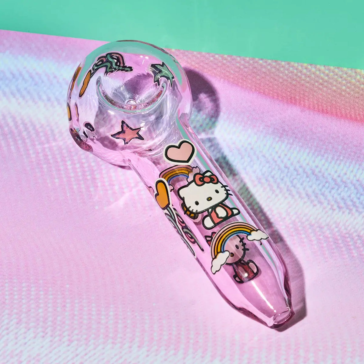 A pink glass water pipe decorated with Hello Kitty and rainbow illustrations, resting on a pink iridescent surface with a mint green background.