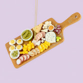 Wooden charcuterie board ornament with assorted meats, cheeses, and fruits on a purple background