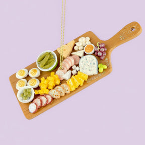 Wooden charcuterie board ornament with assorted meats, cheeses, and fruits on a purple background