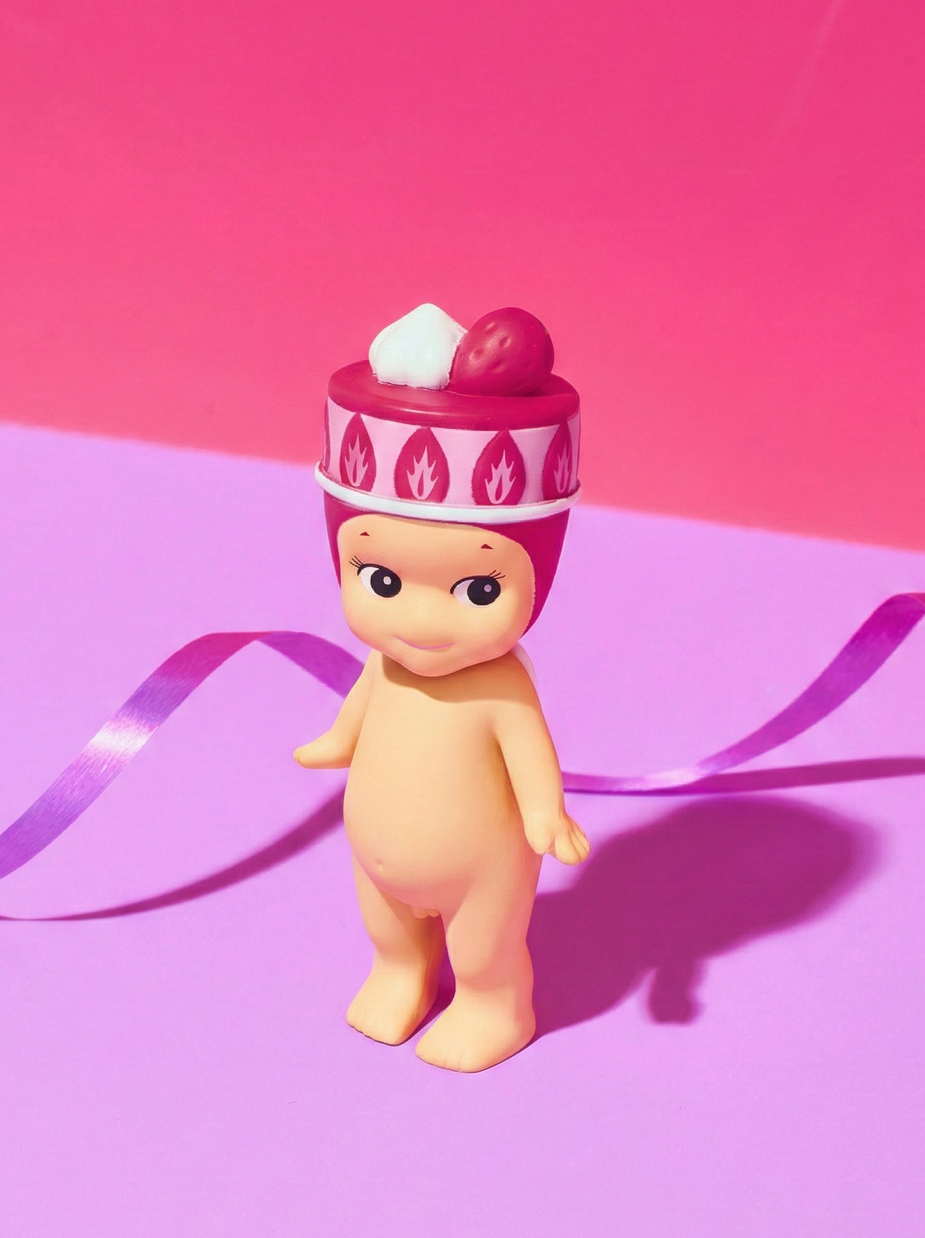 Toy figure with a red hat on a pink cake hat on a two tone background